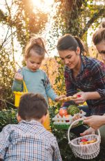 Zwei Personen halten jeweils ein kleines Osternest in den Händen, während zwei Kinder im Garten nach Ostereiern suchen.