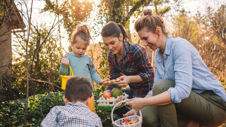 Zwei Personen halten jeweils ein kleines Osternest in den Händen, während zwei Kinder im Garten nach Ostereiern suchen.