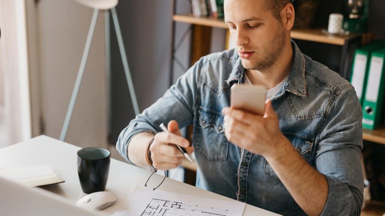 Raumplaner, Richte mit diesen Apps dein Zuhause ein Eine Person sitzt an einem Schreibtisch. Vor ihr liegt ein Zettel mit einem Grundriss einer Immobilie. In der einen Hand hält sie einen Stift, in der anderen ein Smartphone.