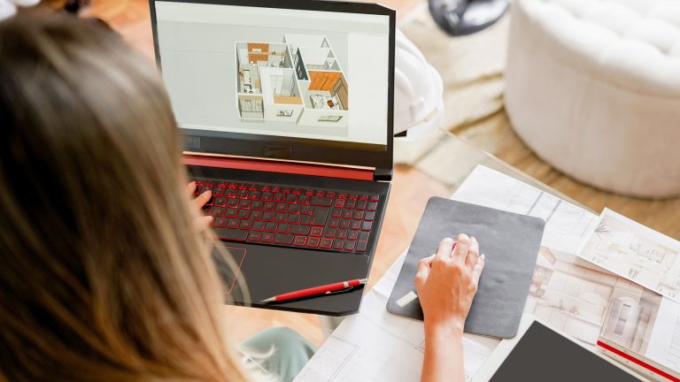 Raumplaner, Richte mit diesen Apps dein Zuhause ein Eine Person sitzt an einem Glas-Schreibtisch. Auf dem Laptop vor ihr ist ein 3D-Grundriss eines Raumes zu sehen. Daneben liegen jede Menge Zettel mit Zeichnungen.