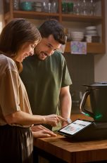 Zwei Personen stehen in der Küche an einem Tisch, auf dem der neue TM7 steht. Eine der beiden Personen bedient diesen per Touchscreen.