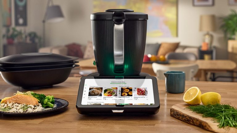 Thermomix TM7 Der TM7 steht auf einer hölzernen Arbeitsplatte. Daneben steht ein Teller mit einer fertigen Reis-Speise sowie ein Holz-Schneidbrett mit Dill und einer aufgeschnittenen Zitrone.