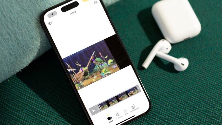 Videos bearbeiten, Apps zum Schneiden, Bearbeiten und Co. Ein iPhone liegt auf einem grünen Untergrund. Darauf die geöffnete Fotos-App mit einem Video im Bearbeitungsmodus. Daneben liegen AirPods und ein zugehöriges Case.