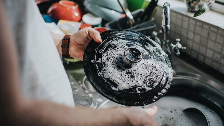 Wasserverbrauch Spülmaschine, Hand vs. Maschine Eine Person hält einen Bräter-Deckel in der Hand, der noch von Schaum bedeckt ist. Im Hintergrund ist weiteres Geschirr in der Spüle zu sehen.