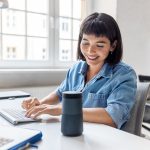 Eine Person sitzt an einem Schreibtisch, tippt auf einer Tastatur und lehnt sich in Richtung eines Echo-Lautsprechers von Amazon.