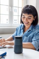Eine Person sitzt an einem Schreibtisch, tippt auf einer Tastatur und lehnt sich in Richtung eines Echo-Lautsprechers von Amazon.