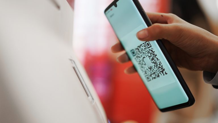 QR-Code erstellen, Mit diesen Apps und Services klappt es Eine Person hält einen QR-Code auf einem Smartphone an einen QR-Scanner an einem Terminal.