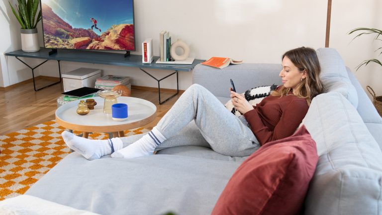 Eine Person liegt auf einer Couch und schaut auf ihr Smartphone. Im Hintergrund ist neben einem Fernseher eine Fritz!Box 6690 Cable zu sehen.