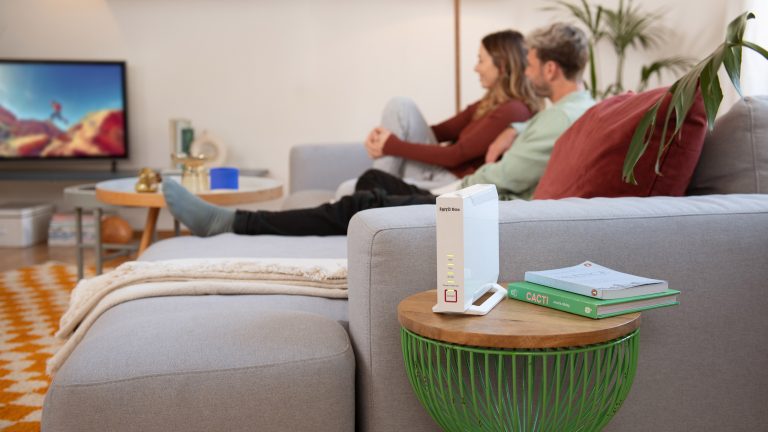 Neben einer Couch, auf der zwei Personen sitzen, steht eine Fritz!Box 4060.