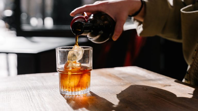 Auf einem Tisch steht ein Glas mit Eiswürfeln. Es ist die Hand einer Person zu sehen, die ein bronzefarbenes Getränk einschenkt.