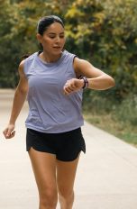 Eine Person in Trainingsklamotten läuft auf einem Weg durch einen Park. Sie blickt auf die Smartwatch an ihrem linken Handgelenk.