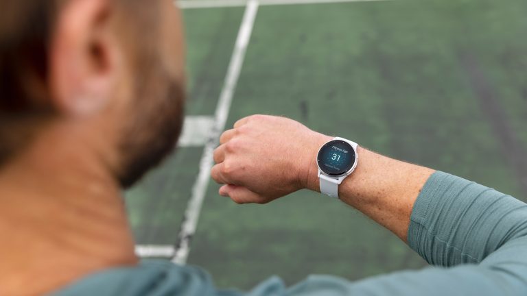 Garmin Connect synchronisiert nicht, Probleme & Lösungen Blick über die Schulter einer Person: Sie steht am Rand eines Sportplatzes und blickt auf die Smartwatch an ihrem rechten Handgelenk.