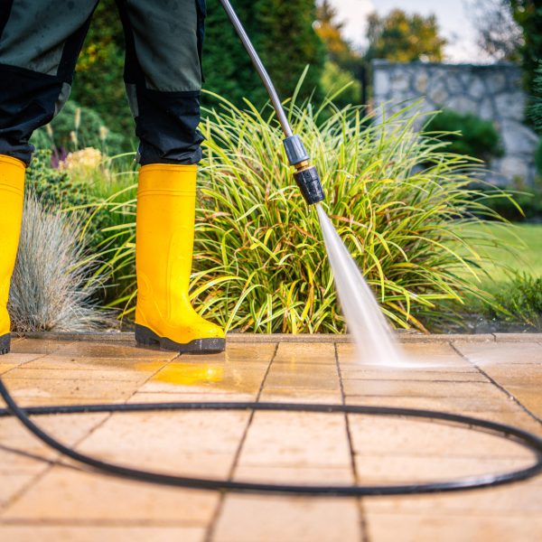 Es sind die Beine einer Person zu sehen, die Gummistiefel trägt. Mit einem Hochdruckreiniger säubert diese die Terrasse in einem Garten.