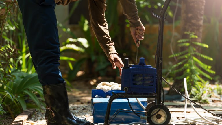 Hochdruckreiniger reparieren, Häufige Probleme und einfache Lösungen Eine Person steht in einem Garten vor einem geöffneten Hochdruckreiniger und säubert Verstopfungen mit einem Schraubenzieher.