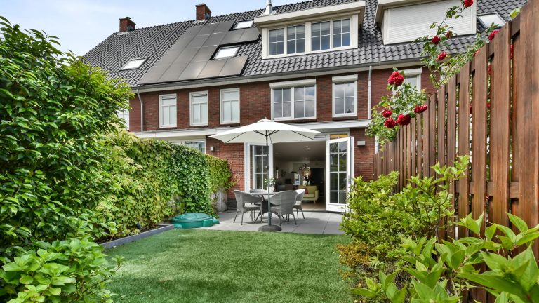 Mähroboter für den kleinen Garten, Darauf kommt es an Blick in den kleinen Garten eines Reihenhauses, der von einem Holzzaun umschlossen ist. Auf der Veranda stehen Gartenmöbel, die Flügeltüren zum Hausinneren sind geöffnet.