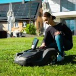 Eine Person kniet hinter einem Mähroboter mit einem geöffneten Gehäuse.