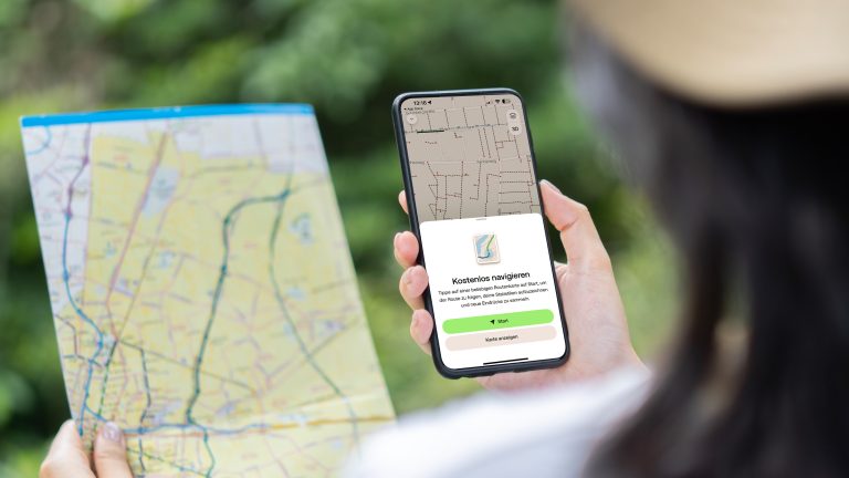 Auf dem richtigen Weg, Wandern mit Wander-Apps Blick über die Schulter einer Person, die in der linken Hand eine Landkarte hält und in der rechten ein Smartphone mit der geöffneten AllTrails-App.