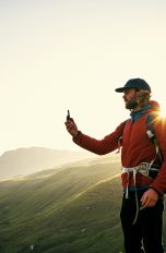 Eine Person steht in Wanderkleidung an einer felsigen Küste und hält ein Smartphone vor sich in der Hand.