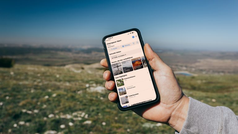 Auf dem richtigen Weg, Wandern mit Wander-Apps Im Vordergrund steht die Hand einer Person, die ein Smartphone mit der geöffneten Bergfex-App in der Hand hält. Im Hintergrund ist eine hügelige Naturlandschaft zu sehen.