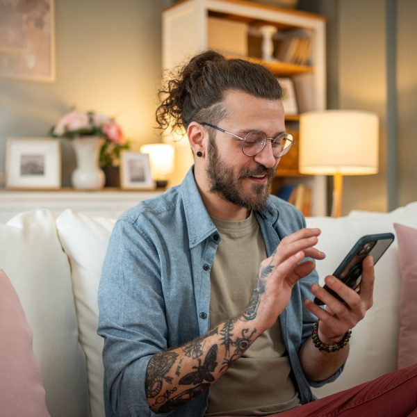 Eine Person mit Bart, langen Haaren und Tattoos sitzt im Wohnzimmer auf der Couch und scrollt auf ihrem Smartphone vor sich in der Hand.