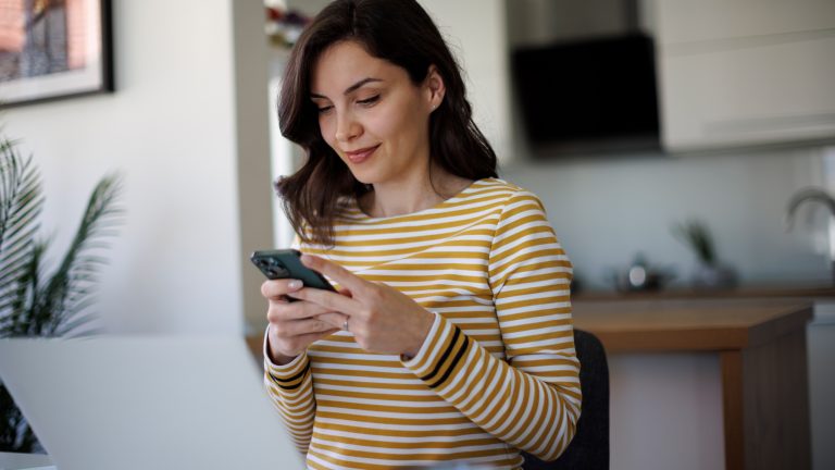 Whatsapp-Alternativen, 6 sichere Messenger im Vergleich Eine Person sitzt in ihrer Wohnung an einem Tisch. Vor ihr steht ein Laptop und sie hält ein Smartphone in beiden Händen vor sich.