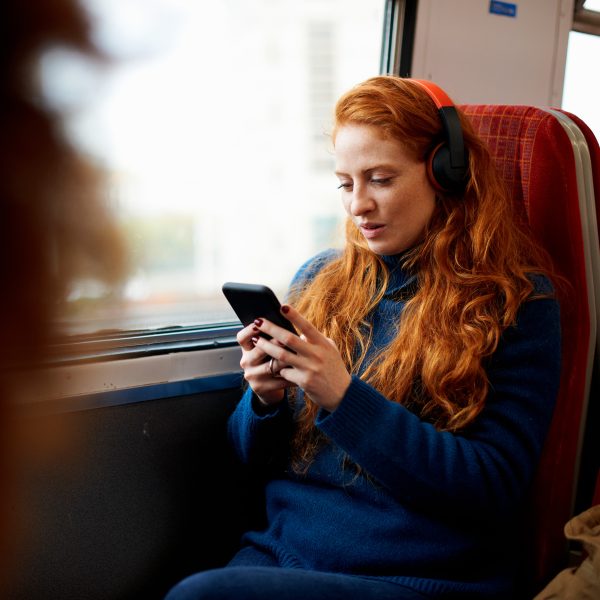 Eine Person sitzt in einem Zug am Fenster, trägt Overear-Kopfhörer und schaut auf ihr Smartphone.