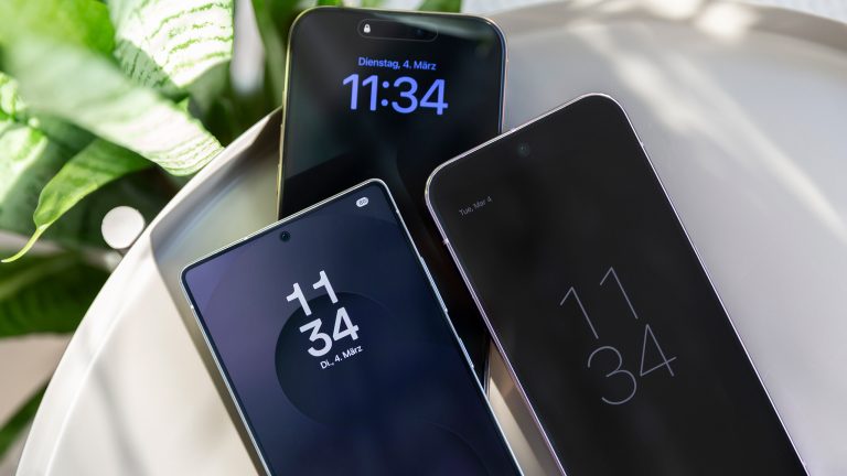 5G Handys, Was können sie? Ein iPhone 16 Pro Max, ein Samsung Galaxy Ultra und ein Google Pixel 9 Pro XL liegen schräg übereinander auf einem runden Metalltisch. Links oben eine Pflanze im Anschnitt.