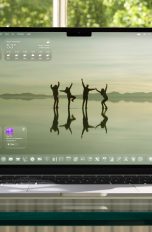 macOS 26 & iPadOS 26, Wer braucht jetzt noch einen Mac? Ein aufgeklapptes MacBook auf einem Glasschreibtisch vor einem Fenster. Auf dem Screen der Startbildschirm unter macOS 26.