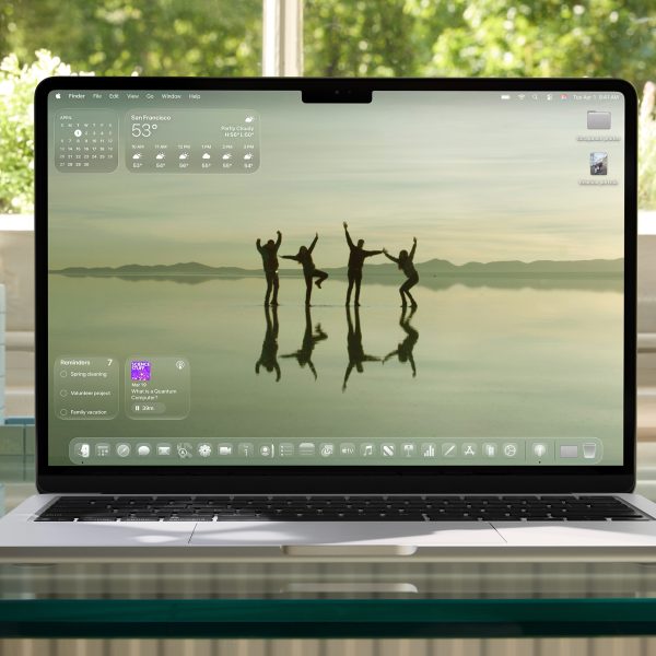 Ein aufgeklapptes MacBook auf einem Glasschreibtisch vor einem Fenster. Auf dem Screen der Startbildschirm unter macOS 26.
