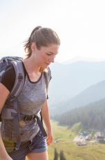Seitansicht einer Person in Jeansshorts und mit Wanderrucksack. Im Hintergrund ist alpine Landschaft zu sehen.