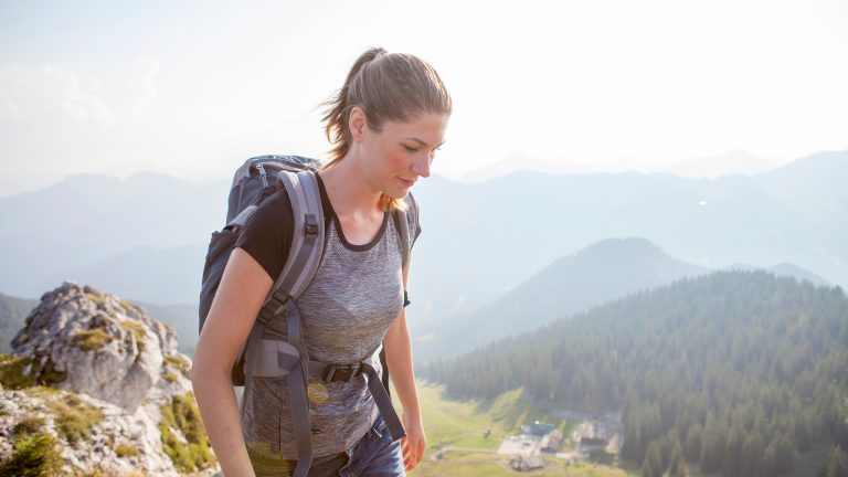 Geschenkidee für Outdoor-Fans, Sechs geniale Technik-Gadgets Seitansicht einer Person in Jeansshorts und mit Wanderrucksack. Im Hintergrund ist alpine Landschaft zu sehen.