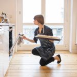 Eine Person öffnet mit einem Backblech in der Hand kniend einen Ofen in einer Küche.