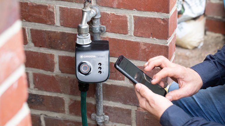 Eine Person kniet neben einem Wasseranschluss, an dem ein Gardena Smart-System angeschlossen ist und bedient ihr Smartphone.