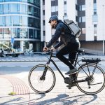 Eine Person auf einem Fahrrad fährt durch eine Stadt. Auf ihrem Kopf ist ein schwarzer Helm zu sehen.
