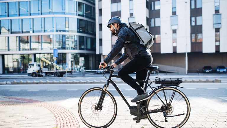 Eine Person auf einem Fahrrad fährt durch eine Stadt. Auf ihrem Kopf ist ein schwarzer Helm zu sehen.