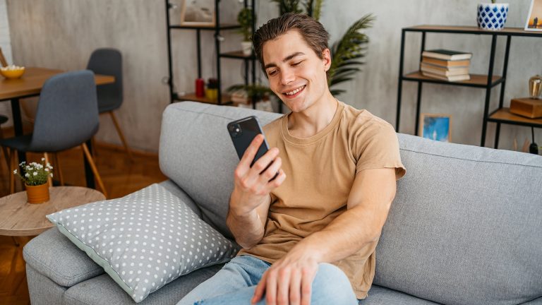 Telefonieren mit Telegram, So gehen Sprach- und Videoanrufe Eine Person sitzt auf einem Sofa in einem Wohnzimmer und blickt auf ihr Smartphone.
