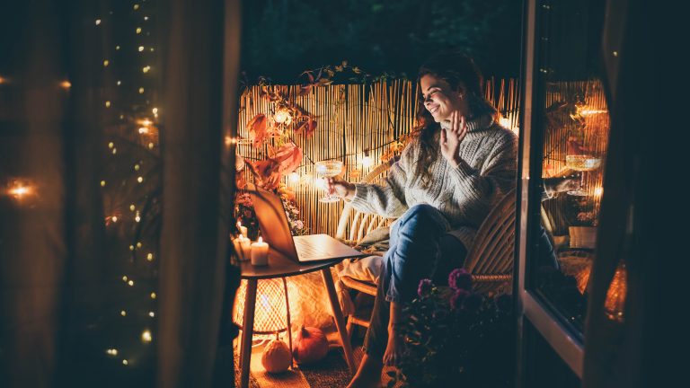 Eine Person sitzt im Dunkeln auf ihrem gemütlich gestalteten Balkon, hält ein Weinglas in der Hand und sieht lächelnd in ihren Laptop. Im Hintergrund ist eine Lichterkette zu sehen.