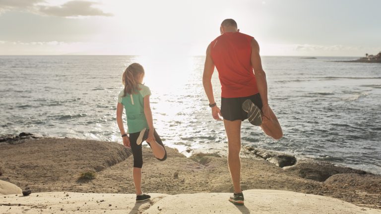 Urlaub mit Kindern, 12 Gadgets für die Kleinen Eine Person und ein Kind stehen auf den Felsen am Meer und machen Dehnungsübungen.