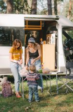 Zwei Personen und ein Kind stehen vor einem Camper auf einem Campingplatz. Auf dem Campingtisch bereiten sie gerade Essen vor.