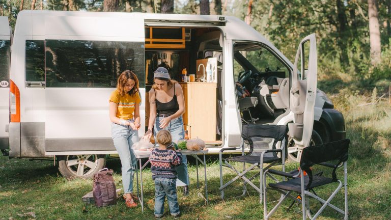 Zwei Personen und ein Kind stehen vor einem Camper auf einem Campingplatz. Auf dem Campingtisch bereiten sie gerade Essen vor.
