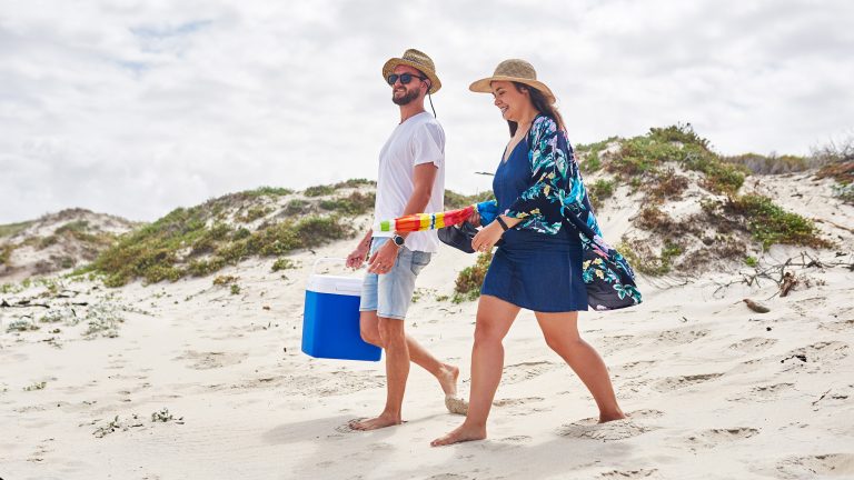 Elektrische Kühlboxen, Modelle, Technik & Tipps für unterwegs Zwei Personen laufen in Strandkleidung über sandige Dünen. In den Händen tragen sie einen Sonnenschirm und eine Kühlbox.