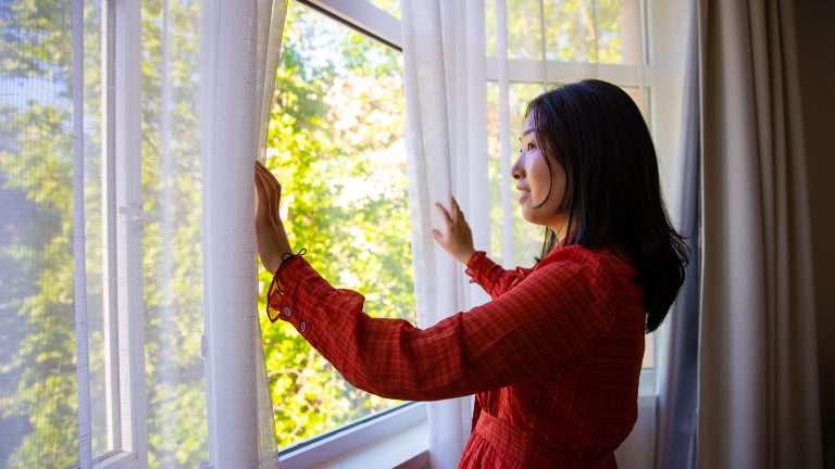 Eine Person steht an einem geöffneten Fenster. Sie öffnet den Vorhang, der vor dem Fenster hängt, mit beiden Händen.