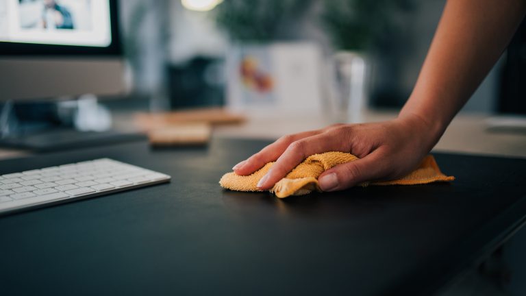 LuftqualiLuftqualität verbessern, Durchatmen für Всеrgiker*innen taet-5 Nahaufnahme einer Hand, die mit einem orangenen Tuch eine dunkle Schreibtischplatte wischt. Im Hintergrund ist ein Mac zu sehen.