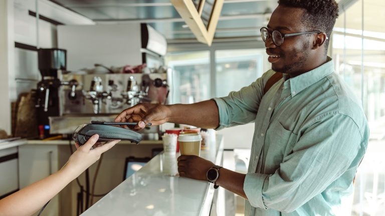 Was ist NFC? So setzt du die Funktechnik ein Eine Person steht am Tresen eines Cafés. In der linken Hand hält sie einen Kaffeebecher, in der rechten das Smartphone, mit dem sie gerade bezahlt.