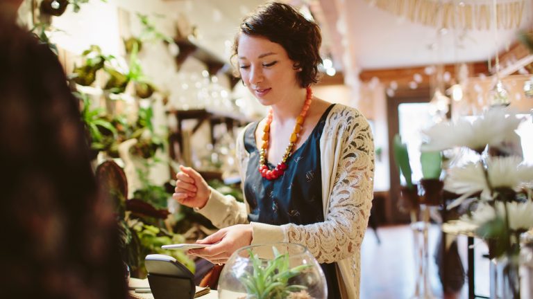 Eine Person steht in einem Blumenladen an der Kasse. Sie hält ihr Smartphone ans Lesegerät, um zu bezahlen.