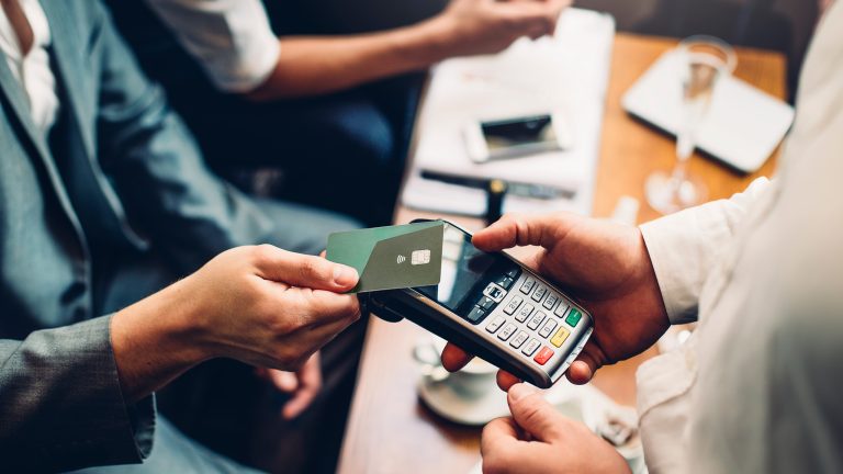 Was ist NFC? So setzt du die Funktechnik ein Ansicht von oben auf eine Person, die an einem Tisch in einem Café sitzt. Sie hält ihre Kreditkarte an ein Kartenlesegerät, das von einer anderen Person in den Händen gehalten wird.