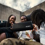 Vier Personen sitzen zusammen und lachen. Eine Person hält einen Bluetooth-Lautsprecher in den Händen.