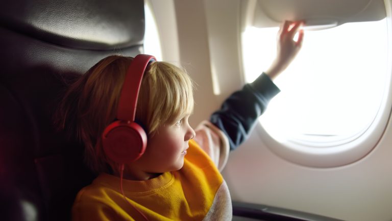 Ein Kind sitzt in einem Flugzeugsitz, trägt Over-Ear-Kopfhörer und schließt gerade die Blende des Flugzeugfensters.