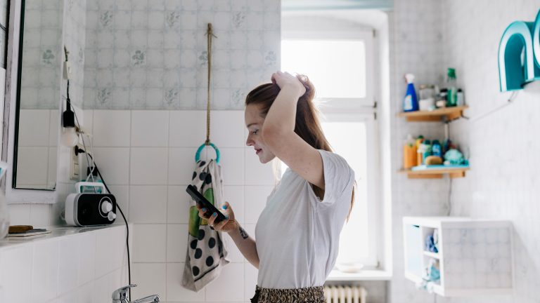 Eine Person steht im Badezimmer vor dem Spiegel. Mit der linken Hand fasst sie sich in die Haare, in der rechten Hand hält sie ein Smartphone.