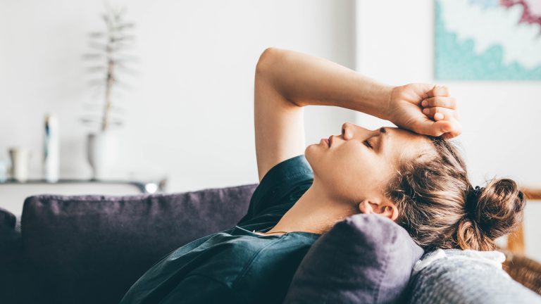 Zyklus-Apps, Wie sie Körper, Gesundheit und Partnerschaft stärken können Eine Person liegt auf einem Sofa. Den Kopf hat sie nach hinten gestreckt, die Augen geschlossen. Eine Hand, die zur Faust geballt ist, liegt auf der Stirn.
